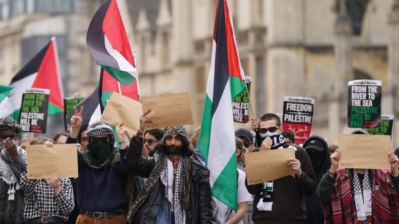 People at a protest wearing masks and scarves, holding brown pieces of paper and green, black and white flags.