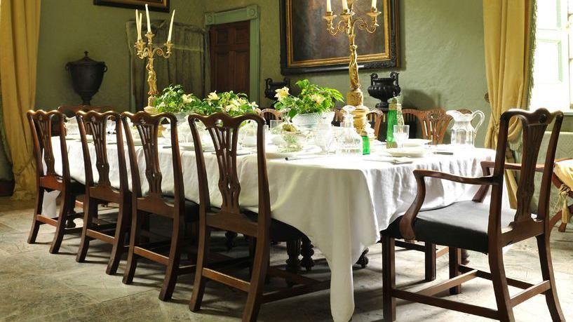 A long dining room table, draped with a white tablecloth and with green plants, gold candlesticks and expensive crockery on top of it. There are 10 wooden chairs around it, and in the background there are gold curtains and a painting on the wall.