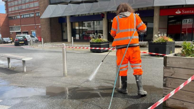 A worker in high-vis orange workwear is using a jet to clean dirt off the pavement near a Farnborough shopping centre. Dark grey water and muck is in the foreground as cleaner paving appears.
