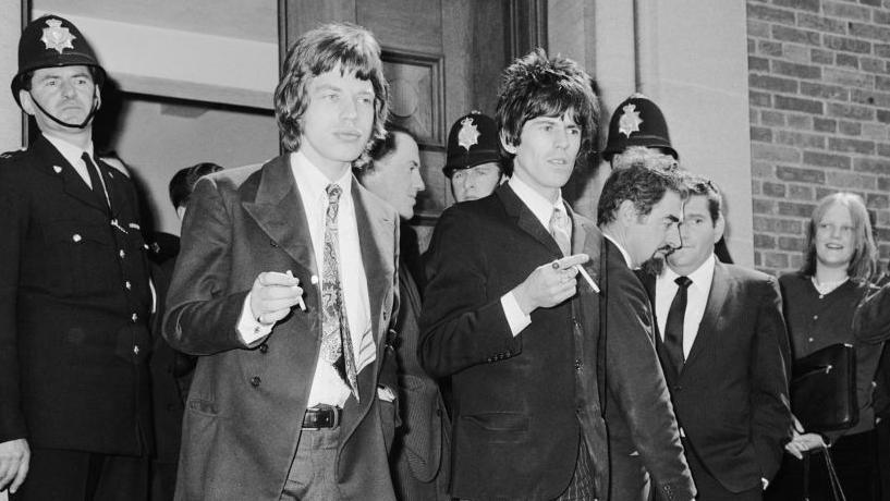 A black and white image of Mick Jagger and Keith Richards in suits, smoking cigarettes, outside the courthouse in Chichester