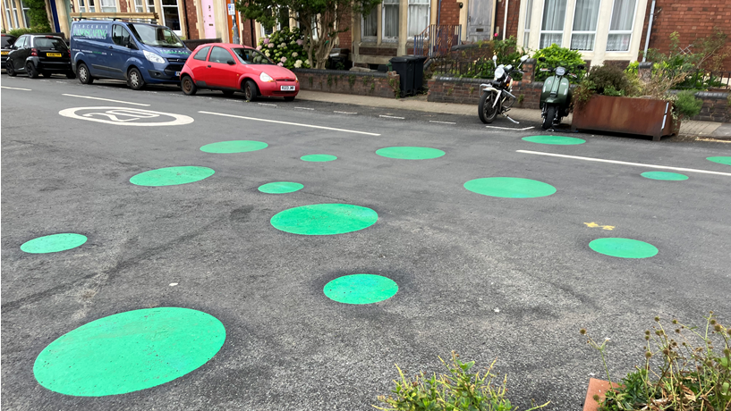 A cluster of large, green spots can be seen painted on a road. A 20 mph speed limit is also painted on the surface. A row of houses can be seen - and several vehicles parked at the kerbside.