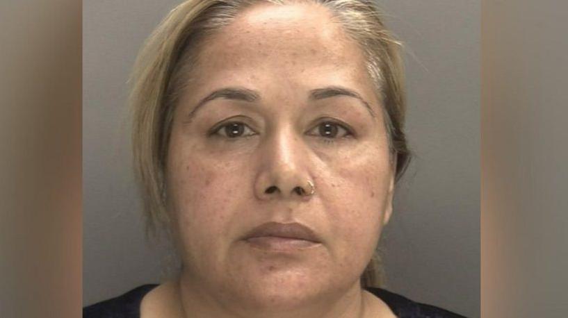 Farzana Kauser with blonde hair in a ponytail and a black top, stood in front of a grey background for her mugshot.