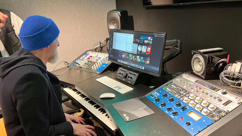 Ian Dench sitting at a mixing desk with mixers and a computer screen with a keyboard under the desk, just visible.
Ian is wearing a black hoody and a blue beanie hat. There are a number of speakers on the desk and soft studio walls.