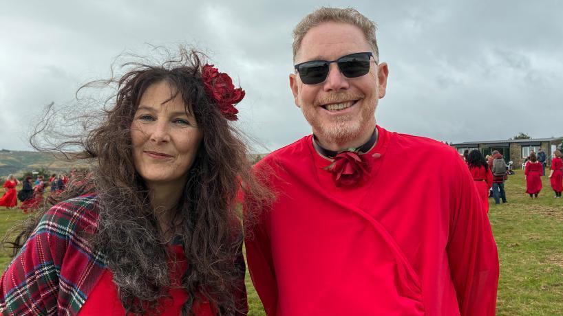 A man and a woman stand in a field, wearing red.