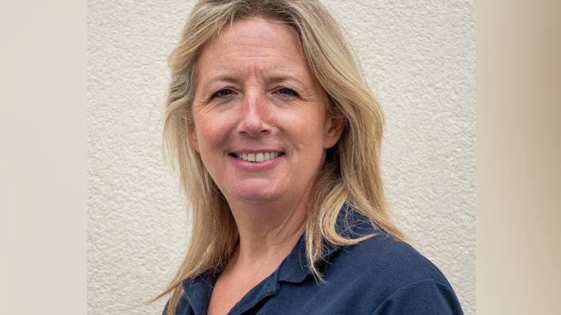 A portrait of Sarah Berrey who has shoulder-length blonde hair and is looking direct into the camera, with a smile and wearing her blue RNLI T-shirt