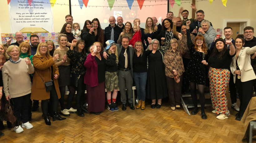 A large group of people in a school hall are looking to the camera. Some have an arm raised in celebration