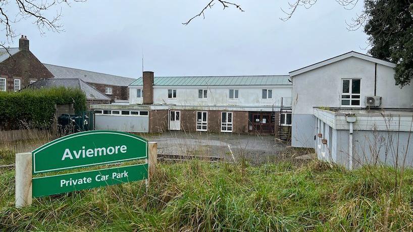 An L-shaped building with a car park in front of it, in the foreground is a grass mound with the Aviemore Private Car Park sign