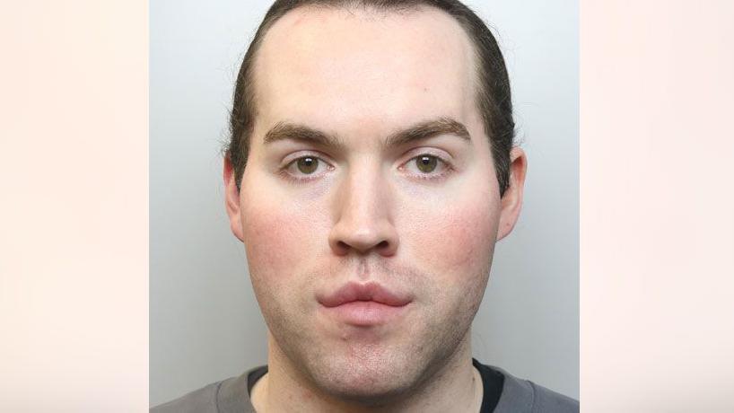 A police mugshot of Jack Watkin. He has long brown hair which is tied back and is wearing a grey top with a dark coloured top underneath.