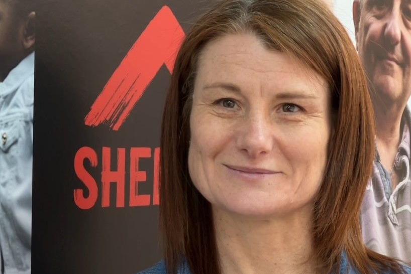 Tracy Guy from housing charity Shelter. She has brown shoulder length hair and brown eyes. She is wearing a blue denim dress. She is pictured standing in front of a poster promoting the charity. 