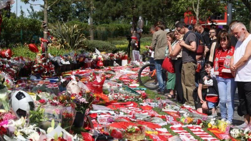 A crowd of fans pay respects at tributes to Jota. One woman has a hand over her mouth. A man in a white vest bows his head to reflect. 