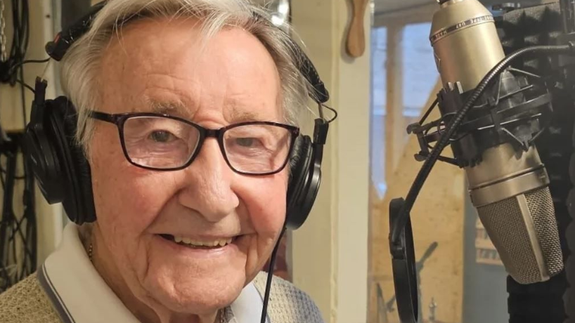 Mr Rayner looking at the camera and smiling. He has brown eyes and white hair. He is wearing headphones and is sitting in front of a microphone.