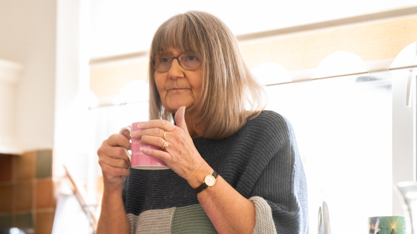 Gill Rich, a 71-year-old woman with a short white hair, leaning in front of her kitchen sink holding a mug while looking out of shot 