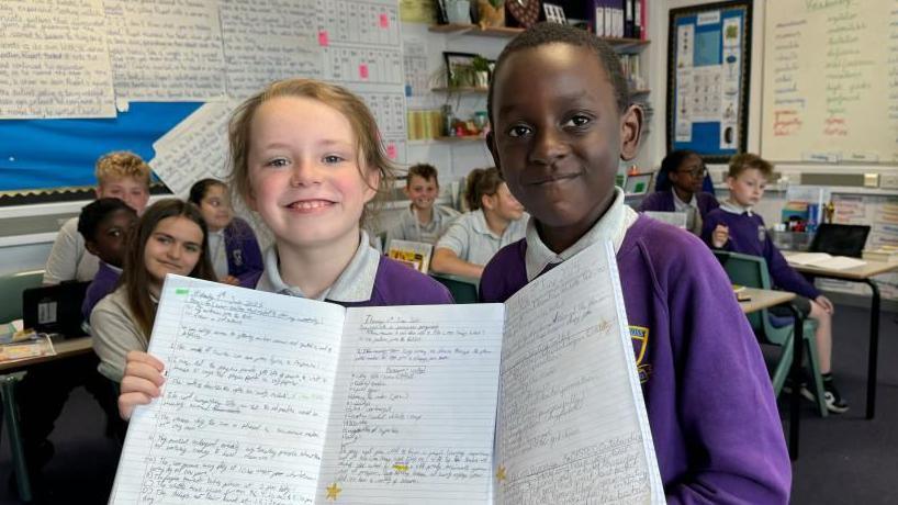 Maxwell has blonde hair, tied back, while Rayleigh has short black hair. They are both wearing white polo shirts and purple jerseys, embroidered with the school logo. They are holding their A4-sized workbooks open. Other children are sitting at desks behind them. School work is displayed on the classroom's walls.