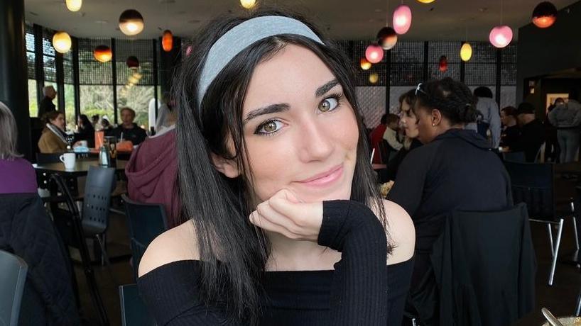 Juliette is smiling at the camera, resting her chin on her hand. She has long, dark straight hair and is wearing a blue headband and black long-sleeved, off-the-shoulder top. In the background is a restaurant with people sitting at tables and coloured lights coming down from the ceiling. 