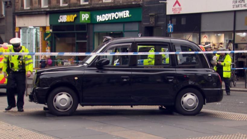 A black taxi sitting on the pavement behind blue and white police tape. Police officers are in the background.