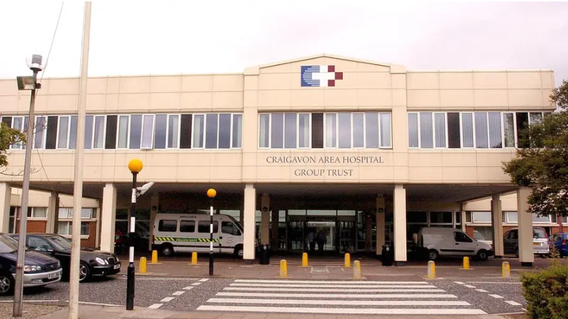 A big off-white hospital facade with a medical crest below the ridge. Parked out the front are ambulances and there is a zebra crossing in foreground, 