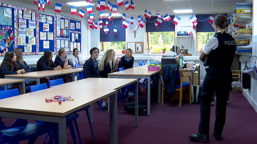 Students in a classroom with two rows of tables, back row has five girls sat across and the front row has three girls. A police woman is standing at the front of the class. The walls are covered in posters and the ceiling has French flag bunting. Burgundy flooring and three windows on the wall furthest away.