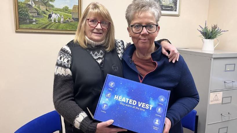 Two women holding a heating pack. Janet has shoulder length blonde hair with red rimmed glasses and is wearing a grey and white jumper with a grey gilet. Jayne, with short grey hair and dark glasses, is wearing a blue zipped jumper. They are posing in an office setting.