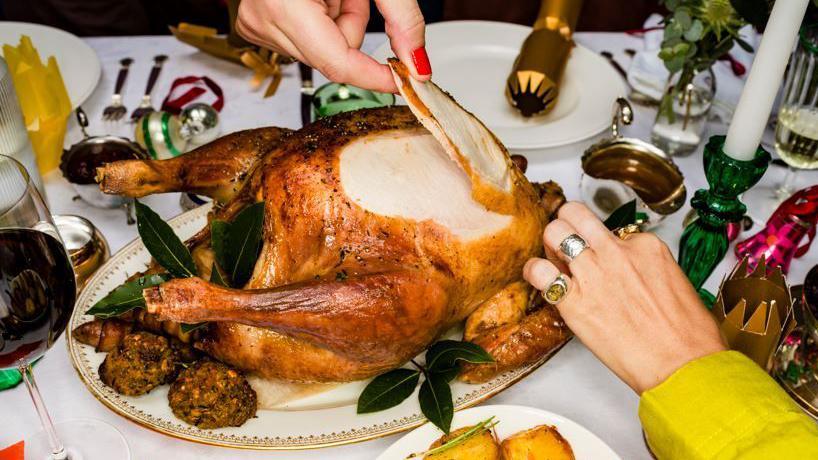 Hands taking slices of meat from a cooked, golden Christmas turkey. Also on the table are vegetables and other trimmings and Christmas crackers  