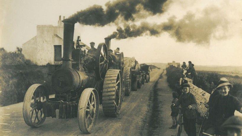 Stream driven engines with plumes of smoke bellowing out of their funnels transporting the whale along a dirt road.