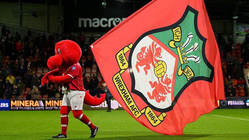 Mascot Wrex the Dragon carries a Wrexham flag across the pitch