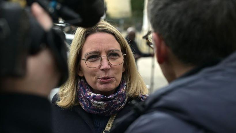 A blonde-haired woman in glasses talks to reporters