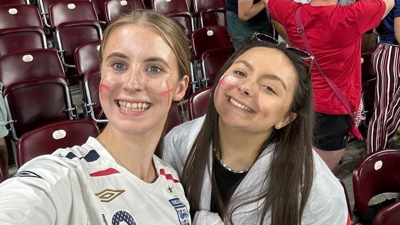 Maisie Fairweather Prescott and her friend Natalie wearing white England shirts with blue insignia and three lions badges in a stadium surrounded by football fans. Natalie has an England flag wrapped round her.