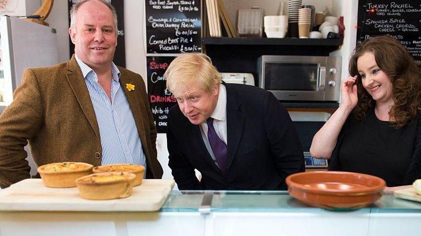 Andrew RT Davies stood to the left of Boris Johnson and Cardiff councillor Jayne Cowan, behind a shop counter. Menus can be seen hung up on the wall behind them. Davies is stood looking at the camera.