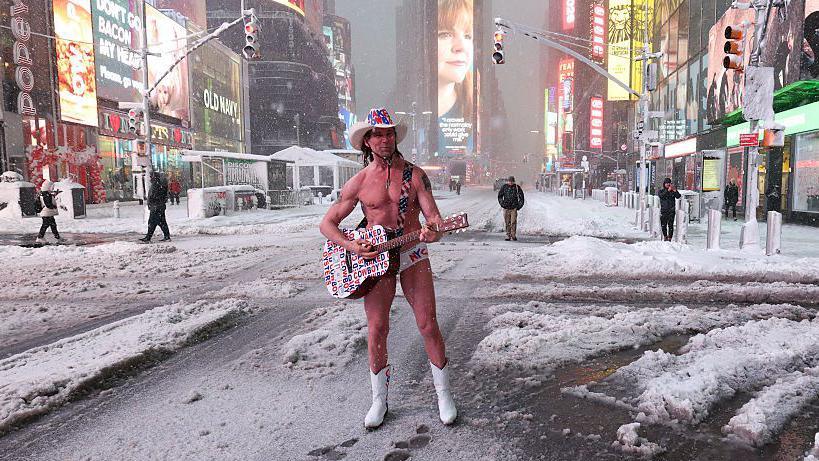 A man wearing only underwear, a cowboy hat and cowboy boots is playing a guitar. Snow is all around him and the lights of the advertisements on the buildings shine brightly. A few other people are walking in the background.
