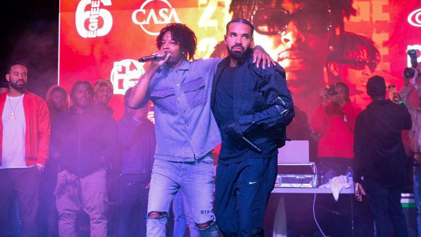 Two men stand on a stage - one holds a microphone to his mouth with one hand, his free hand draped over the other man's shoulder. The second man -dressed in an all-black ensemble including gloves, holds his microphone at waist level as he looks off to the side intently.