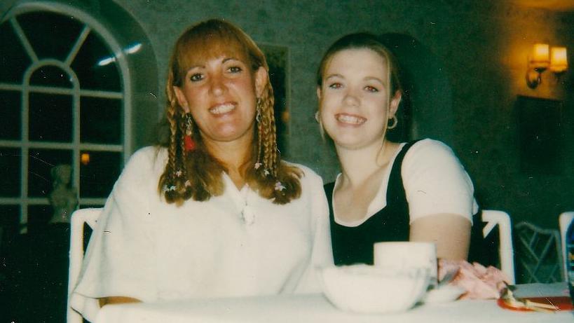 Gemma Collins and her mother, Joan. Both are sitting at a table and smiling at the camera. Joan is wearing a white shirt while Gemma is wearing a white T-shirt with a black top over it