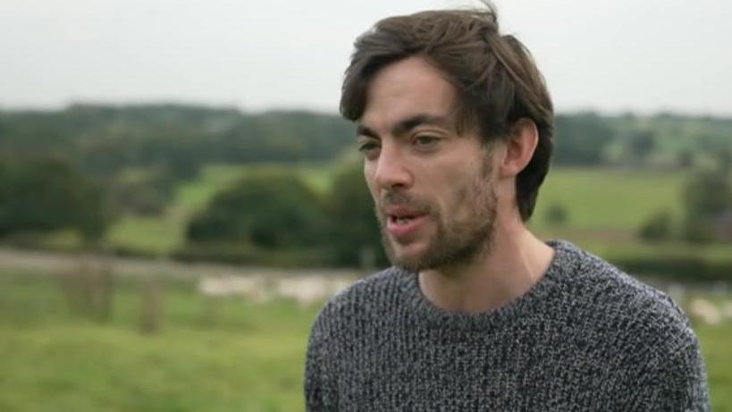 A man with black hair in a grey woolly jumper standing in a field