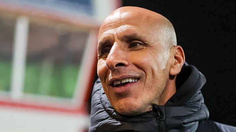 Dino Maamria ahead of a Barrow game, wearing a big puffa coat with the zip done right up to the top