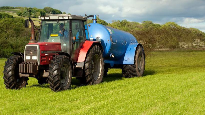 A red tractor pulls a blue trailer as it spreads slurry on a field. 