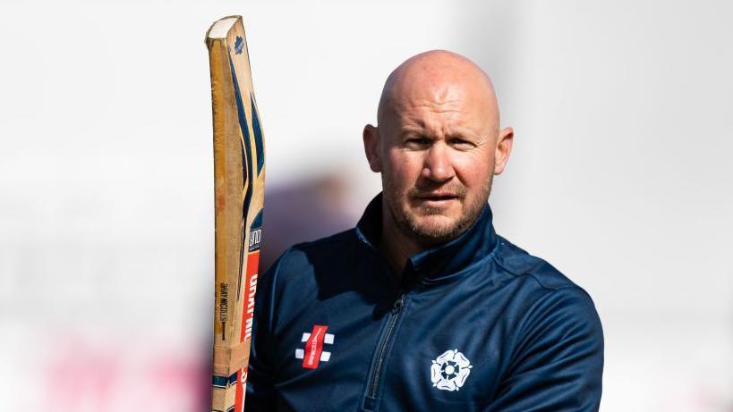 John Sadler holding a cricket bat wearing a Northants tracksuit