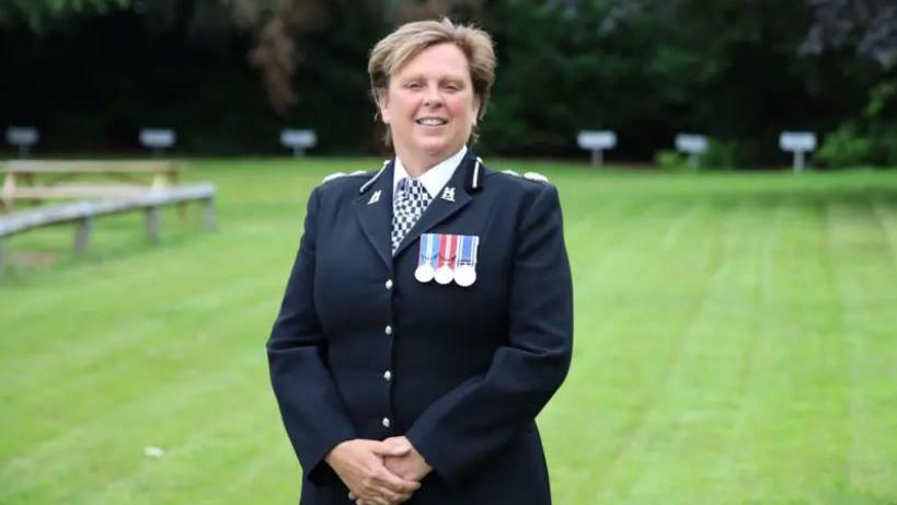 A female police officer looking at the camera. She is wearing a black police uniform, checked tie and three medals. 