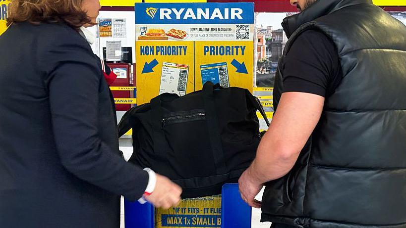 Two unidentified passengers, a man and a woman wearing black, attempt to put a large black bag into a Ryanair bag measurement box. It doesn't look like it fits. The box is in Ryanair's blue and yellow colours, with the words "priority" and "non-priority" with arrows. 