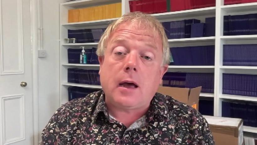 A man with blond hair wearing a brown, purple and cream patterned shirt, sat in a home office with rows of files on white shelving and cardboard boxes behind him.