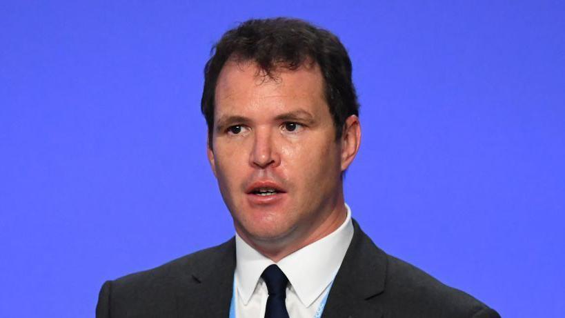 Lee Waters standing in front of a blue background in a black suit and tie