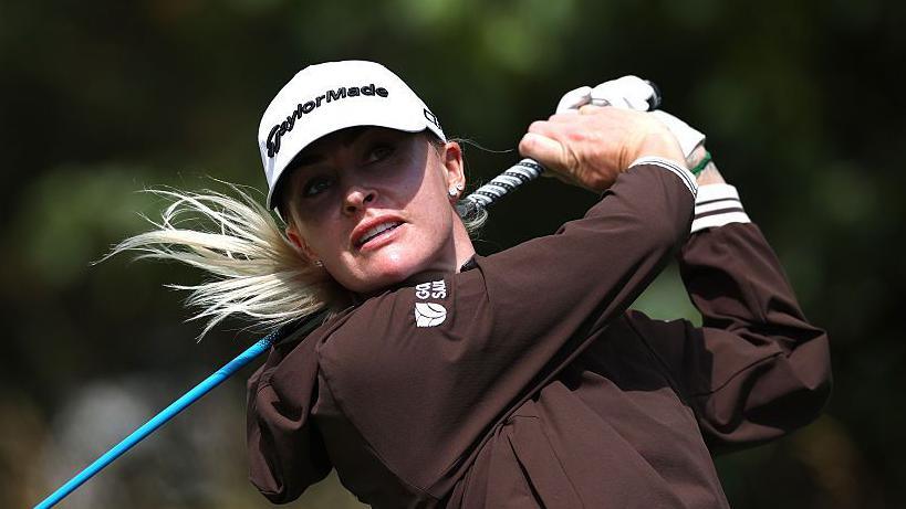 Charley Hull swinging a blue golf club on the course while wearing a white cap, white glove in her left hand and a brown jacket