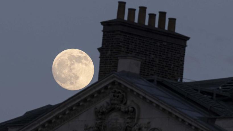 Një hënë e plotë lind në një qiell të kthjellët mbi një çati dhe oxhak. Ndërtesa ka detaje të ndërlikuara dekorative prej guri në çatinë e saj.