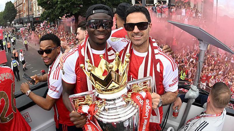 Bukayo Saka and Mikel Arteta, both in sunglasses and red and white team kit, with the Premier League trophy on Arsenal's bus parade