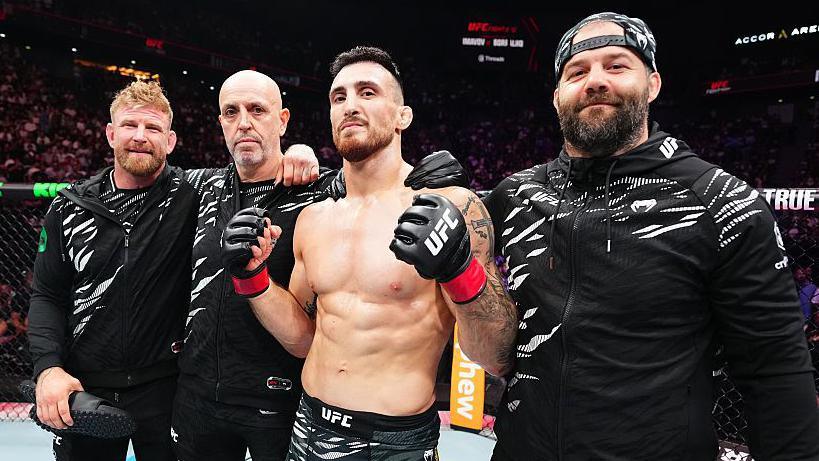 A bare-chested Bukauskas poses smiling with his team in the octagon, including his father Gintas Bukauskas. The three members of his team are dressed all in black outfits, flecked with white