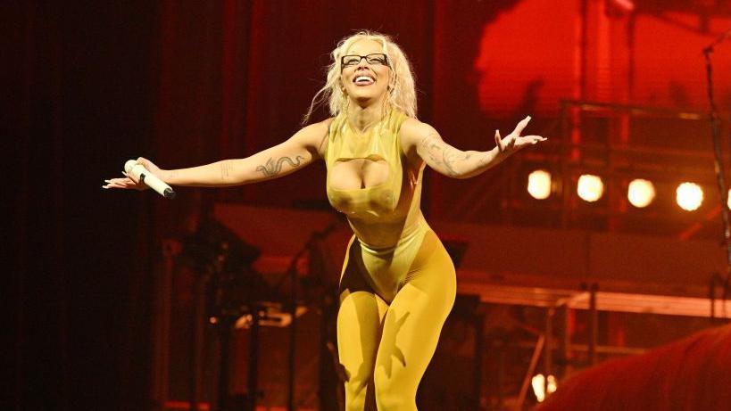 Doja Cat is smiling widely on stage. Her knees are slightly bent and she has her arms stretched out wide. She has a microphone in one hand. Doja Cat is wearing a skin-tight mustard yellow jumpsuit, which has no sleeves and features a keyhoe cut out on the chest. She is wearing black rectangular glasses and has blonde wavy hair which has been tied back into a high ponytail. Behind her is a dark red background with a row of spotlights.