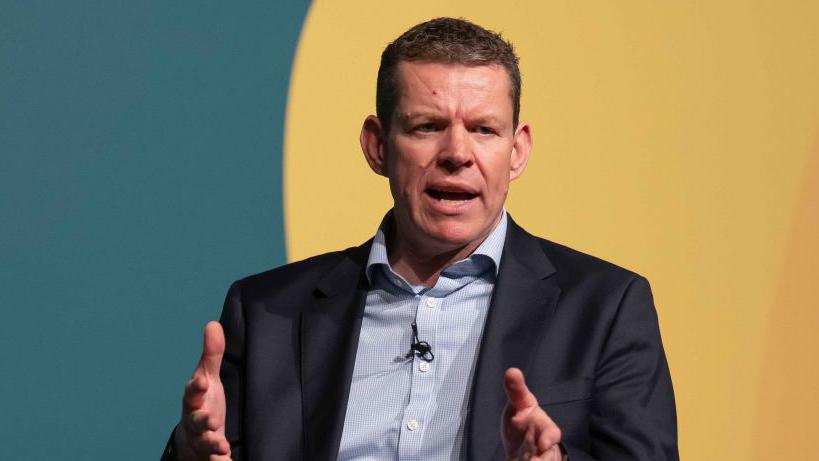Plaid Cymru Leader Rhun ap Iorwerth speaking at a conference. He is sitting in front of a green and yellow wall.