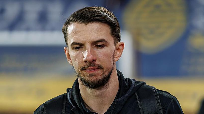 Dan Barlaser walks along a touchline in a black tracksuit with the strap of a bag over his left shoulder