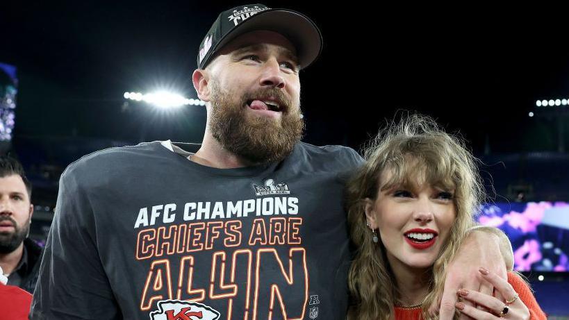 Travis Kelce and Taylor Swift smile to the side of the camera. Taylor wears an orange jumper and has red lipstick and blonde hair. Travis rests his arm on her shoulder and wears a grey AFC champions shirt