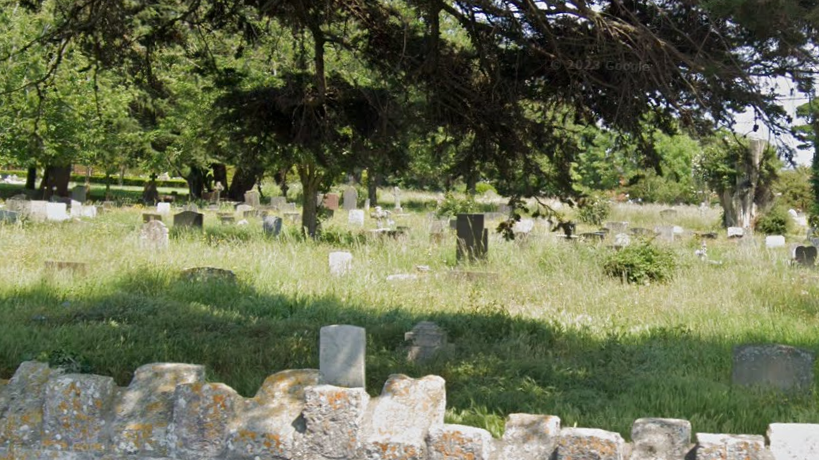 A cemetery with dozens of gravestones. Grass surrounding them has not been cut and is overgrown. There is a stone wall in the foreground