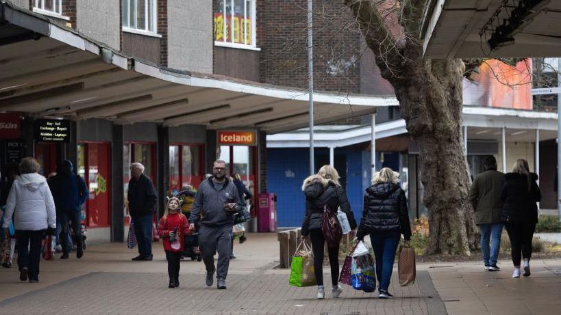 People are walking up and town a parade of shops in Basildon town centre. An Iceland shop front is visible, and a tree in the middle of the pathway