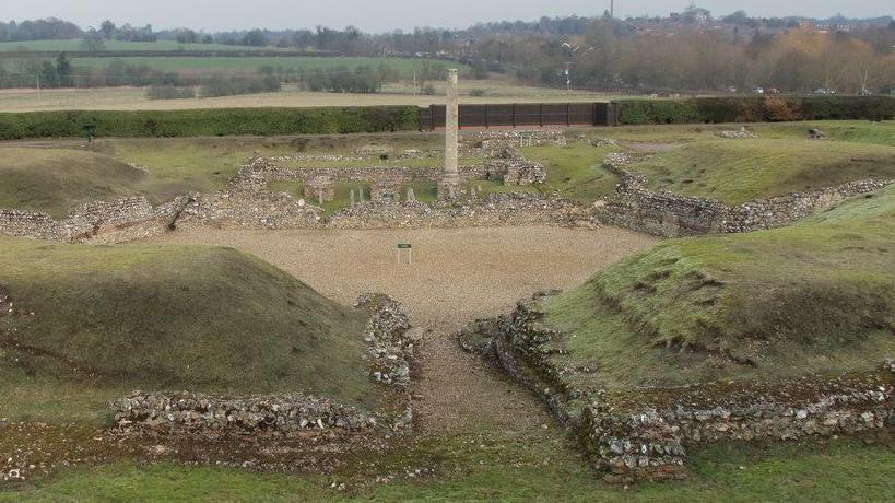 The remains of the Roman Theatre of Verulamium, which was built in about AD140. There are bricks and stones, gravel and grass mounds. Beyond it is countryside.